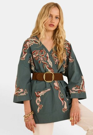 Donna bionda che indossa una giacca verde ricamata con una larga cintura marrone e gioielli d'oro sulla mano sinistra.