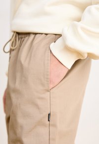 Bežové bavlnené nohavice s elastickým pásom, bočnými vreckami a malou čiernou značkovou nálepkou na ľavom šve. Textúra vyzerá mäkko a hladko.