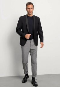Veste de smoking noire avec un revers en satin, portée sur une chemise sombre, associée à un pantalon gris clair et des chaussures habillées noires. Look simple et ajusté.