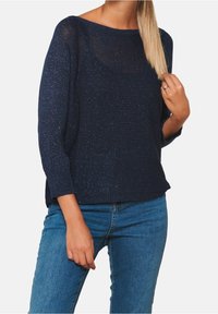 Hafenblau gestrickter Pullover mit kurzen Ärmeln, der über einen lässigen Boot-Ausschnitt verfügt und glänzende Fäden im gesamten Stoff aufweist. Kombiniert mit blauen Jeans.