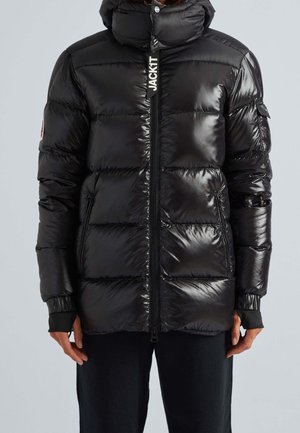 Czarna błyszcząca kurtka puchowa z wysokim kołnierzem, zamkiem błyskawicznym z przodu, bocznymi kieszeniami oraz napisem "JACK IT" na suwaczku, noszona przez stojącą osobę.