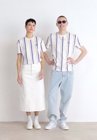 T-shirts blancs rayés avec des lignes verticales bleues, portés avec une jupe blanche et un jean bleu clair. Les deux paires de chaussures sont blanches.