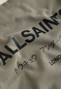 Tissu brun clair texturé avec le texte noir en gras "ALLSAINTS" et un texte plus petit en japonais et en anglais en dessous.