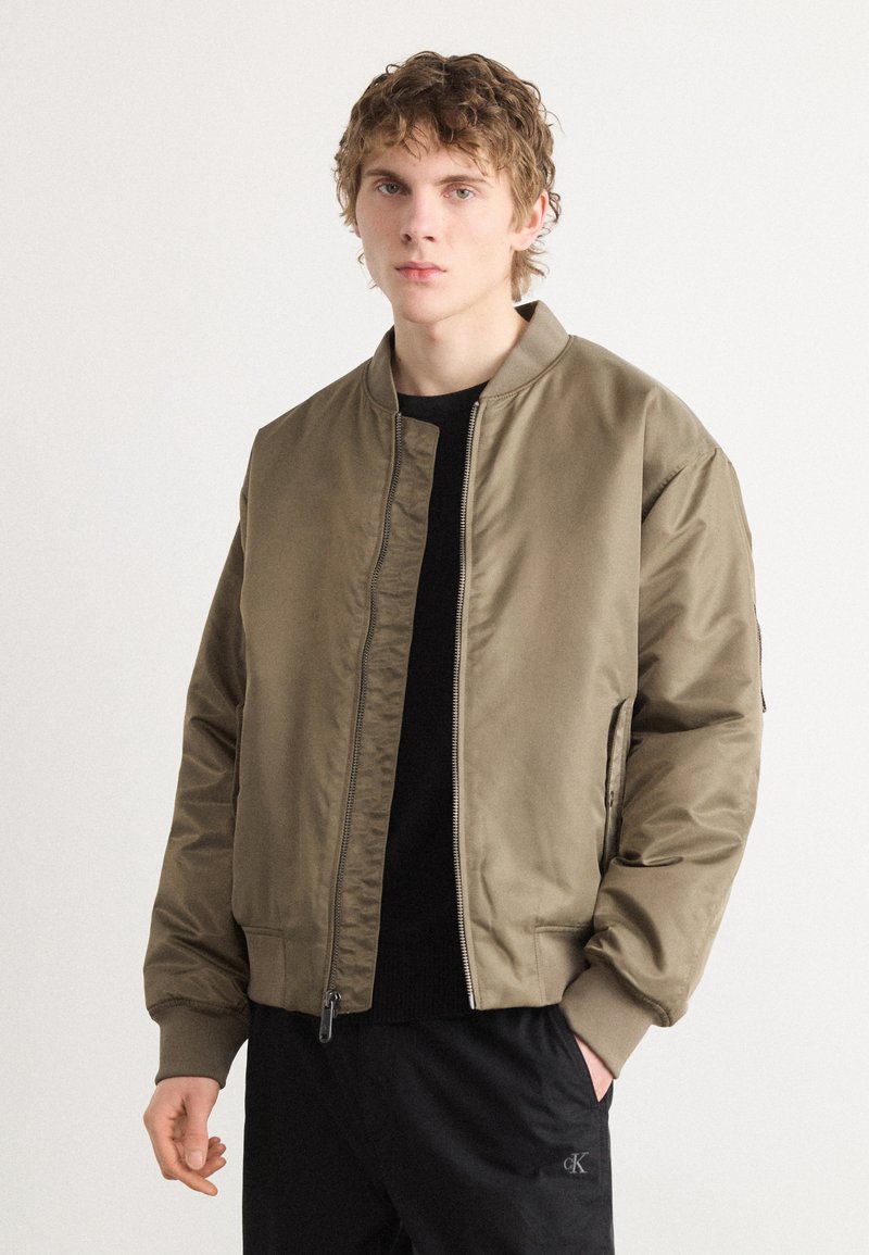 Veste bomber vert olive avec fermeture éclair, col et poignets côtelés, et poches latérales. Portée sur une chemise noire, présentée sur un mannequin.