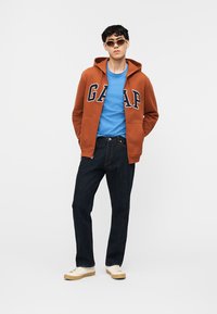 Νέος άνδρας που φοράει πορτοκαλί φούτερ GAP με κουκούλα, μπλε μπλούζα, σκούρο τζιν, λευκά αθλητικά παπούτσια και γυαλιά ηλίου, στέκεται μπροστά σε απλό λευκό φόντο.