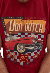 Camiseta roja con un gráfico de coche retro. Los elementos de texto incluyen "Von Dutch Garage" y "AUTO PARTS 24/7". Patrón de cuadros en azul y rojo.
