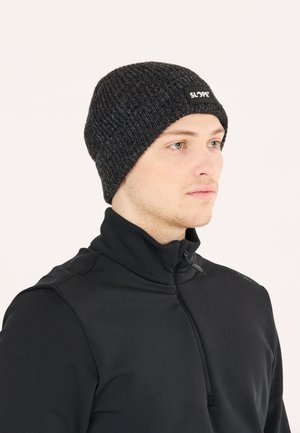 Black knit beanie with a ribbed texture and a small rectangular logo label. Worn with a black zip-up collar jacket.