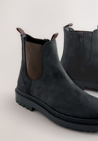 Paar schwarze Stiefeletten mit braunen elastischen Seiteneinsätzen, Zuglaschen, robusten Sohlen und seitlichen Reißverschlüssen auf beigem Hintergrund.