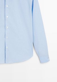 Chemise boutonnée bleu clair à manches longues, en tissu texturé, avec un ourlet arrondi et des boutons blancs.