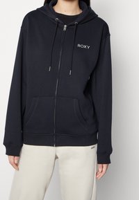 Sweat à capuche zippé bleu marine en tissu doux, avec poches avant, capuche à cordon de serrage et logo blanc sur la poitrine. Porté avec un pantalon de survêtement beige clair.