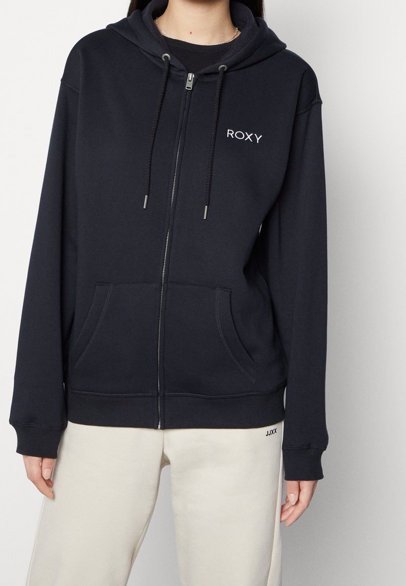 Sweat à capuche zippé bleu marine en tissu doux, avec poches avant, capuche à cordon de serrage et logo blanc sur la poitrine. Porté avec un pantalon de survêtement beige clair.