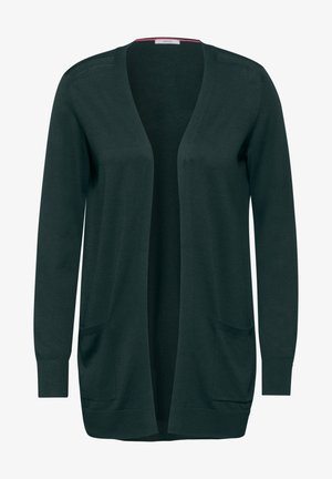 Femme portant un cardigan vert foncé ouvert sur le devant, un t-shirt blanc, un jean bleu large avec revers, et des baskets blanches, debout devant un fond gris.