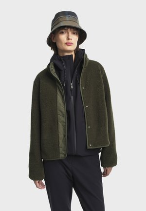 Zöld polárdzsekit viselő személy, fekete cipzáros kapucnis pulóver alatt, és kockás kalapban, egy sima világos háttér előtt áll.