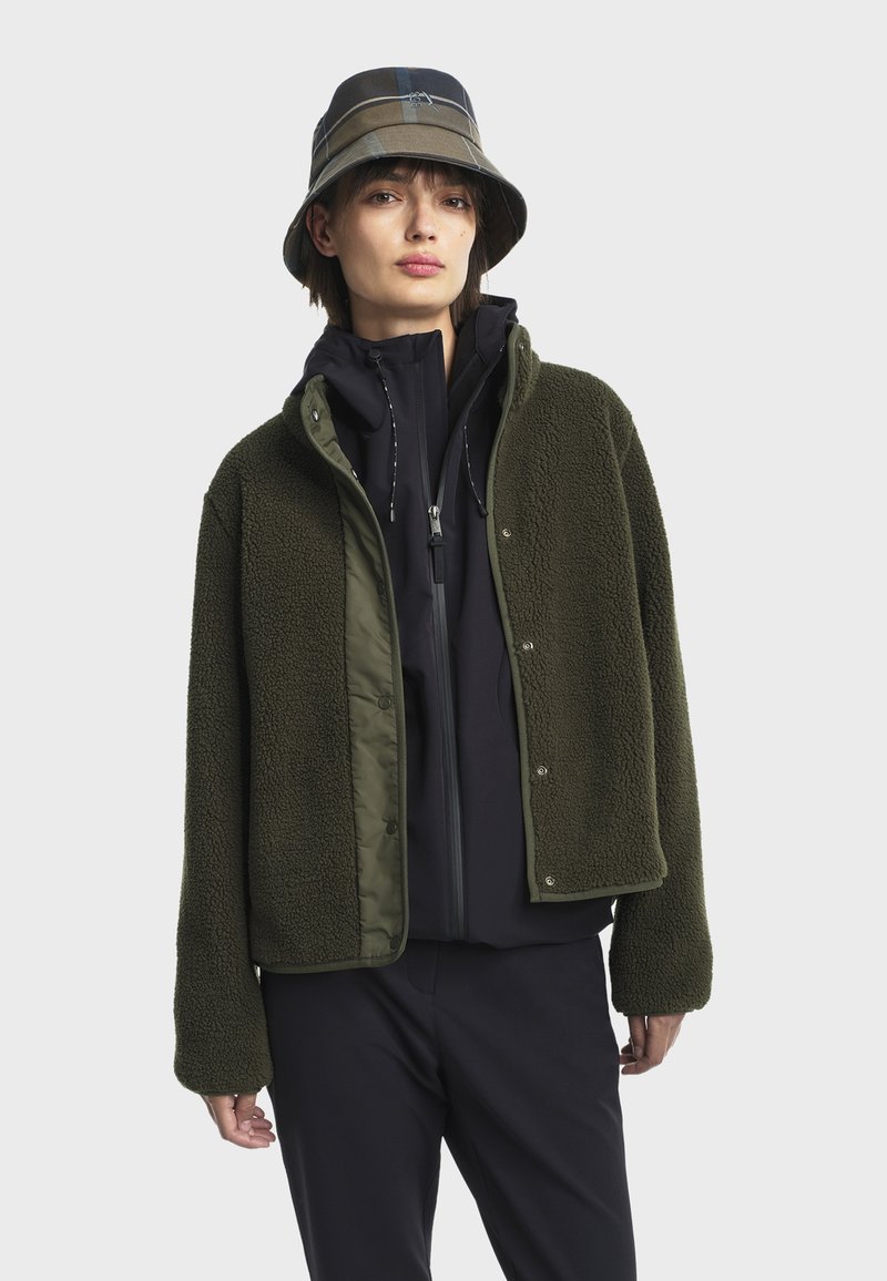 Osoba v zelené fleecové bundě přes černé zipové mikině a s kostkovaným kloboukem, stojící na pozadí v jednobarevném světlém prostředí.