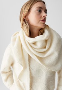 Crème-kleurige, zachte, gebreide sjaal om de hals gedrapeerd. De stof lijkt pluizig met een gladde textuur en subtiele glans.