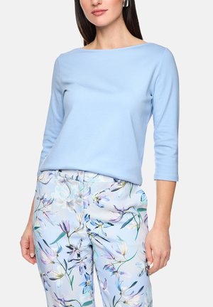 Frau mit hellblauem Dreiviertelarmshirt und hoch taillierter Hose mit pastellfarbenem Blumenmuster vor schlichtem Hintergrund.
