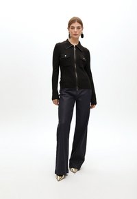 Top negro con relieve y cremallera, con dos bolsillos frontales, combinado con pantalones de mezclilla oscuros de campana y tacones dorados metálicos puntiagudos.