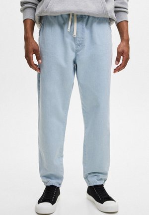 Mężczyzna w jasnoniebieskich dżinsowych spodniach typu jogger, szarej bluzie i czarnych trampkach z białą podeszwą, stojący na jednolitym białym tle.