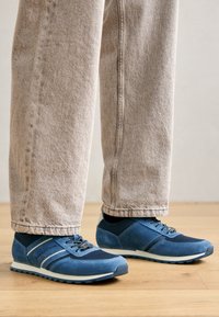 Tênis esportivos azuis com uma mistura de materiais em camurça e malha, solado de borracha, design de cadarço e detalhes brancos contrastantes.
