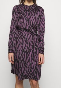Lila och svart klänning med långa ärmar, som har en texturerad djurtryck, rund halsringning och ett knytbälte i midjan.