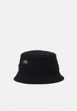Chapeau bob noir avec un petit logo crocodile vert brodé sur le côté, présenté sur un fond uni gris clair.