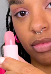 Rosa leppestift i en rosa tube med en avrundet tupp. Hånd som holder tuben med en lys rosa nyanse, plassert mot en nøytral bakgrunn.