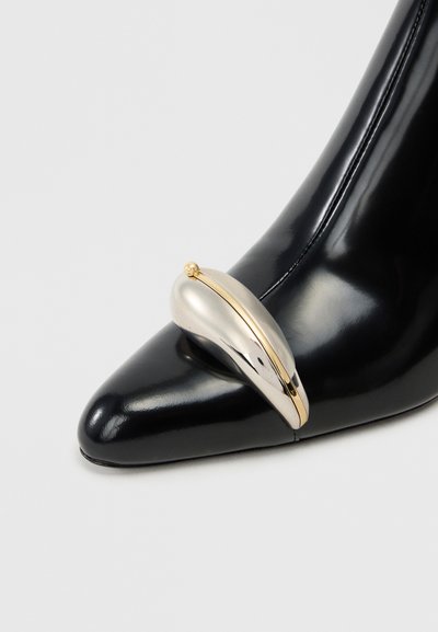 Black leather high heel with pointed toe, featuring a silver and gold sculptural accent at the front. Smooth texture, polished finish.