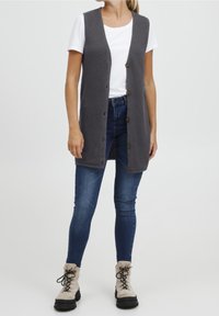 Grau gestrickte lange Weste mit strukturiertem Muster, mit V-Ausschnitt und fünf Knöpfen, getragen über einem weißen T-Shirt und blauen Jeans.