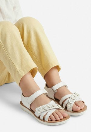 Füße, die weiße Sandalen mit Blumenverzierungen und verstellbaren Riemen tragen, kombiniert mit hellgelben Hosen vor einem einfarbigen Hintergrund.