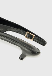 Czarny skórzany sandał na obcasie z odkrytymi piętami, o spiczastym nosku, eleganckim designie, cienkim pasku i małym złotym zapięciu. W zestawie wysoki obcas typu stiletto.