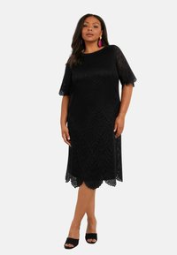 Černé krajkové šaty v těsném střihu s krátkými, zvlněnými rukávy, zdobené vlnovým vzorem a zvlněným lemem, spárované s černými lodičkami na odhalených prstech.
