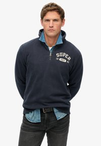 Dunkelblauer Fleece-Pullover mit hohem Kragen und halbem Reißverschluss, mit cremefarbenem Text "SUPER 1985" auf der Brust, über einem Jeanshemd getragen.