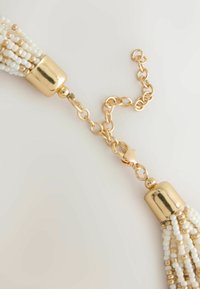 Gold chain necklace clasp connecting two tassels made of white and gold beads on a light background.