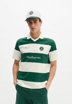 Junger Mann trägt grünes und weiß gestreiftes Poloshirt und passende Kappe, Hände in den Taschen, vor schlichtem weißem Hintergrund stehend.