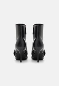 Botines de cuero negro con tacones de aguja altos, que presentan detalles texturizados y un cierre con cremallera decorativa con borla en la parte trasera.