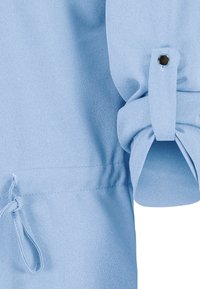 Vaaleansininen paita, jossa on teksturoitu kangas, ominaisuutena gathered-hihansuut ja nappikiinnitys hihassa. Kiristysnauha helman kohdalla.