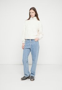 Biely texturovaný pulóver s vysokým golierom, skombinovaný s modrými džínsami s priamymi nohavicami a pruhmi a sivými teniskami. Jednoduchý, moderný dizajn.