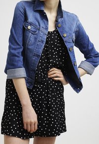 Veste en denim bleue avec un devant à boutons et des poches poitrine, portée par-dessus une robe noire à pois. Dispose de manches retroussées et d'une silhouette ajustée.