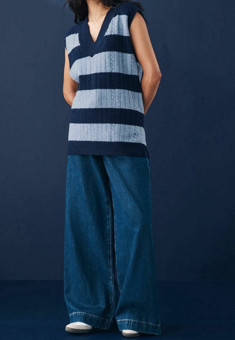 Femme portant un gilet sans manches à col en V avec de larges rayures bleu marine et bleu clair et un jean large bleu foncé ample.