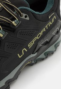 Melnas kalnu pastaigu kurpes ar teksturētu virsū, ar dzeltenu logotipu, GORE-TEX uzlīmi un izturīgu saķeres zoli pelēkā un melnā krāsā.
