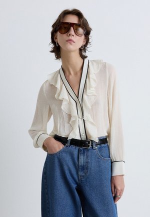 Femme portant de grandes lunettes de soleil marron, un chemisier blanc à volants avec une bordure noire, rentré dans un jean bleu taille haute avec une ceinture noire.