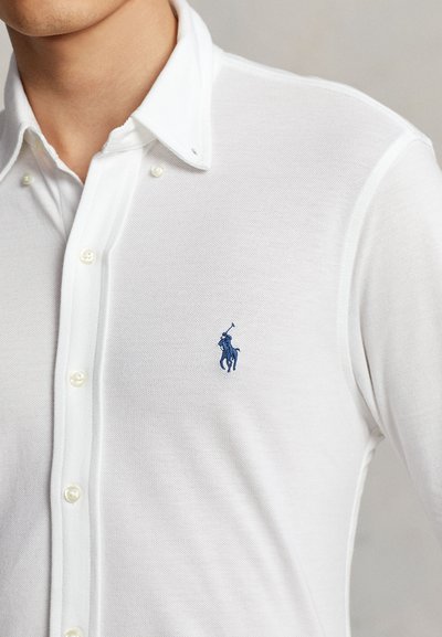Gros plan d'une personne portant une chemise blanche à boutons avec un logo brodé bleu d'un joueur de polo sur la poitrine.