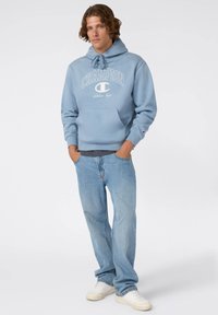 Felpa blu chiaro con cappuccio a cordino e logo "Champion" bianco. Abbinata a jeans blu sbiaditi e sneakers bianche. Vestibilità casual.