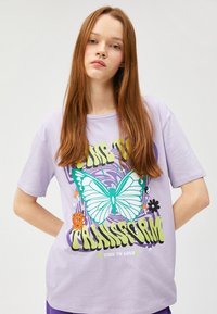 Camiseta lila de corte holgado con un diseño gráfico de una mariposa, un sol y texto que dice "ES HORA DE TRANSFORMARSE" en verde y negro.