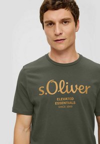 Olivgrünes Baumwoll-T-Shirt mit rundem Ausschnitt und kurzen Ärmeln, mit goldgedrucktem Text "s.Oliver ELEVATED ESSENTIALS SINCE 1969."