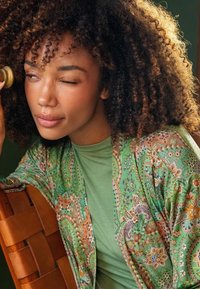 Green patterned kimono with floral and paisley designs, worn over a light green top. Curly hair, soft texture, and relaxed fit. Winking expression.