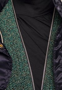 Gilet vert et noir en tricot superposé sur un haut noir, avec une fermeture éclair en argent et des accents en tissu bleu foncé brillant.