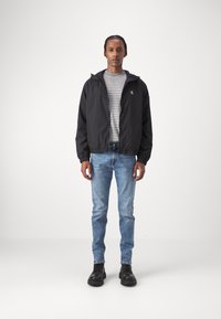 Giacca leggera nera, camicia a righe grigie e bianche, jeans slim-fit blu e stivali neri, in piedi davanti a uno sfondo neutro.