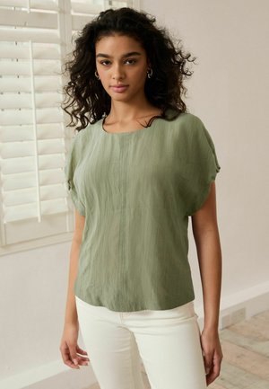 Blusa verde claro con un acabado texturizado, mangas cortas y un corte holgado, combinada con pantalones blancos. Cuello redondeado y sencillo.