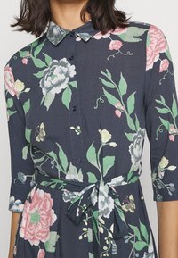 Blommig klänning med mörk marin bakgrund, korta ärmar, knappstängning framtill och ett uppdraget midja med ett matchande bälte, prydd med färgglada blommor.